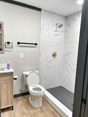 A bathroom with a toilet, a shower with a hexagonal tile pattern, a wooden cabinet, and a handrail.