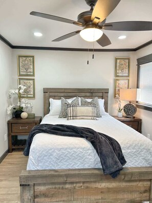 A bedroom with a bed, a ceiling fan, framed artwork, and a nightstand with a lamp.