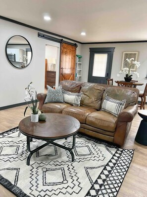 A living room with a brown leather sofa, a round coffee table, a patterned rug, a mirror, and a dining area with a table and chairs.