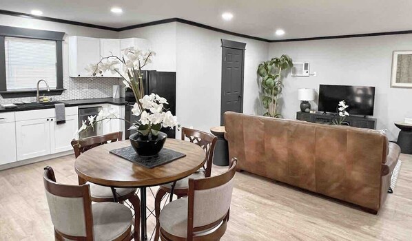 A modern kitchen with white cabinets, a dining table with chairs, a sofa, and a TV in the living area.