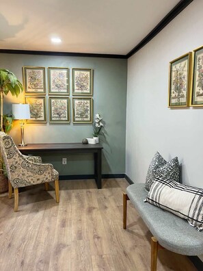 A room with a desk, two chairs, a sofa, a lamp, and framed artwork on the wall.
