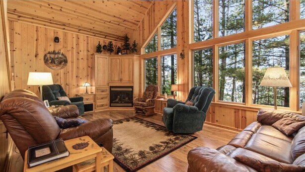 A log cabin interior with a fireplace, leather furniture, and large windows offering a forest view. - Lakefront Whitefish Chain Retreat - Sleeps 15 - Pontoon - Fall Getaway & Rates (Crosslake)