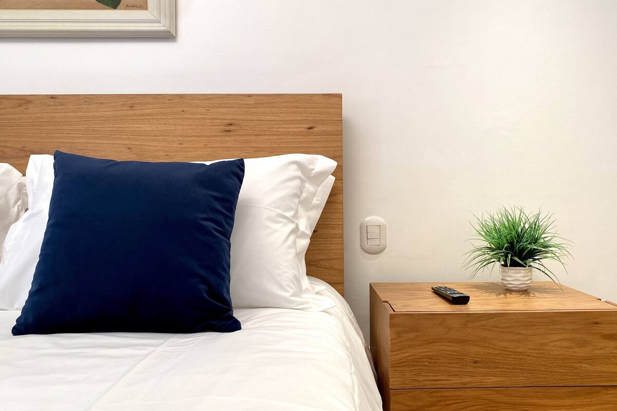 A neatly made bed with white and navy blue pillows, a wooden nightstand with a remote control and a potted plant, and a light switch on the wall.