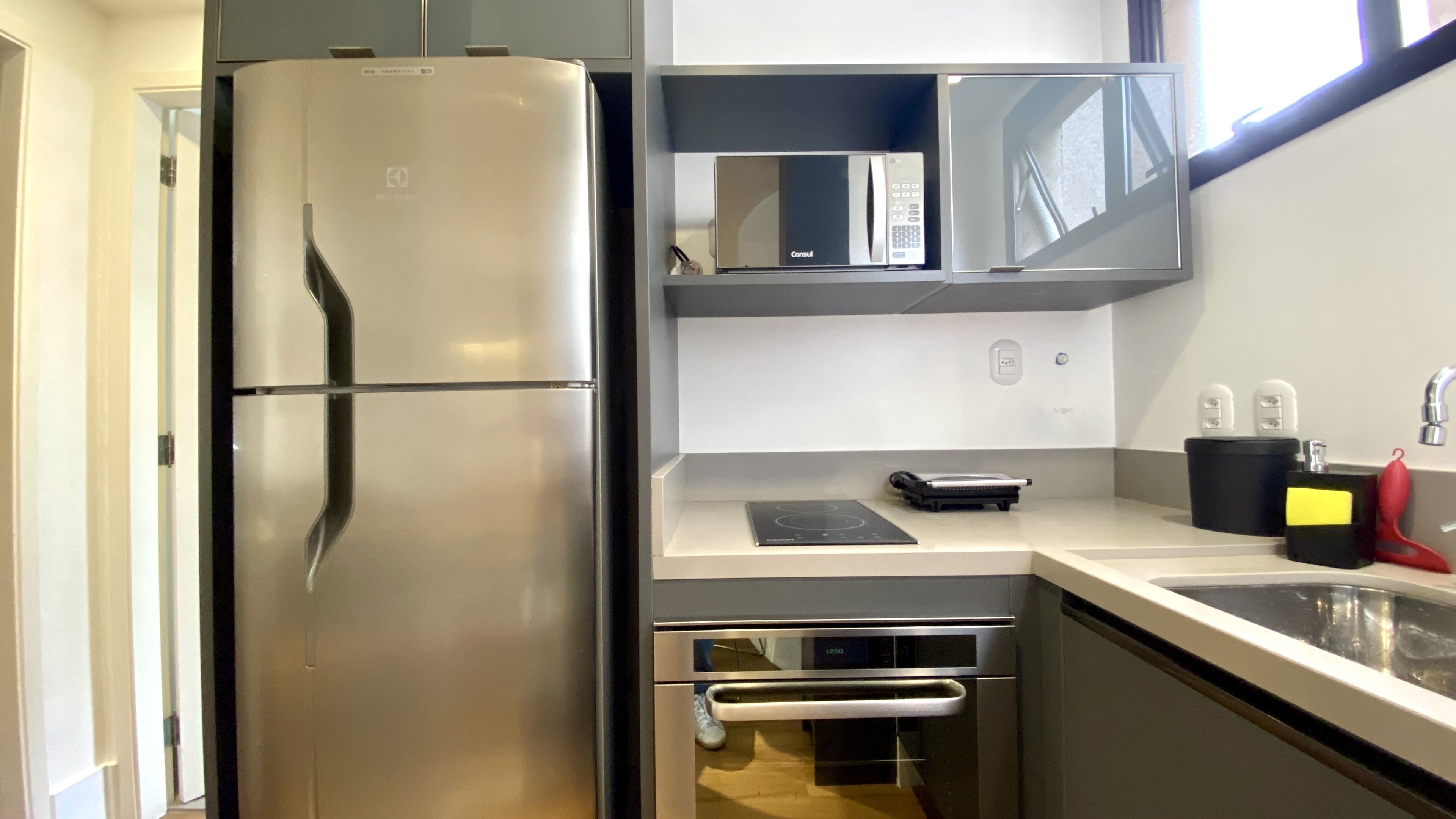 A modern kitchen with a stainless steel refrigerator, a microwave, and a built-in oven. There is a small countertop with a kettle and a red object.