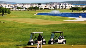 Ein Golfplatz mit zwei Golftrolleys, einem Teich und einer Reihe von Häusern im Hintergrund.