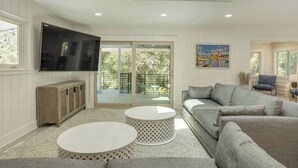A modern living room with a flat-screen TV, a grey sofa, two round white tables, and a balcony with glass doors. - Luxury Beach House - Group/Family/Dog Friendly (Hilton Head Island)