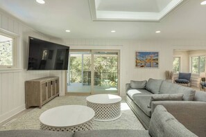 A modern living room with a flat-screen TV, a grey sofa, two round white tables, and a balcony with glass doors.