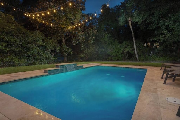 A rectangular swimming pool with a tiled edge, surrounded by a patio area with a table and chairs.
