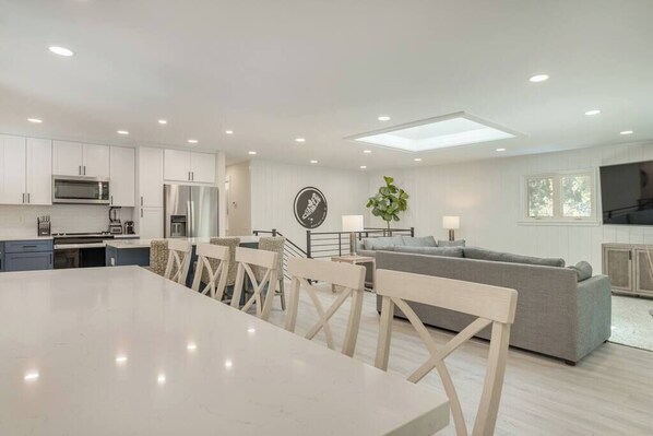 A modern kitchen with white cabinets, a dining table, and chairs. There is a gray sofa and a small plant in the living area.