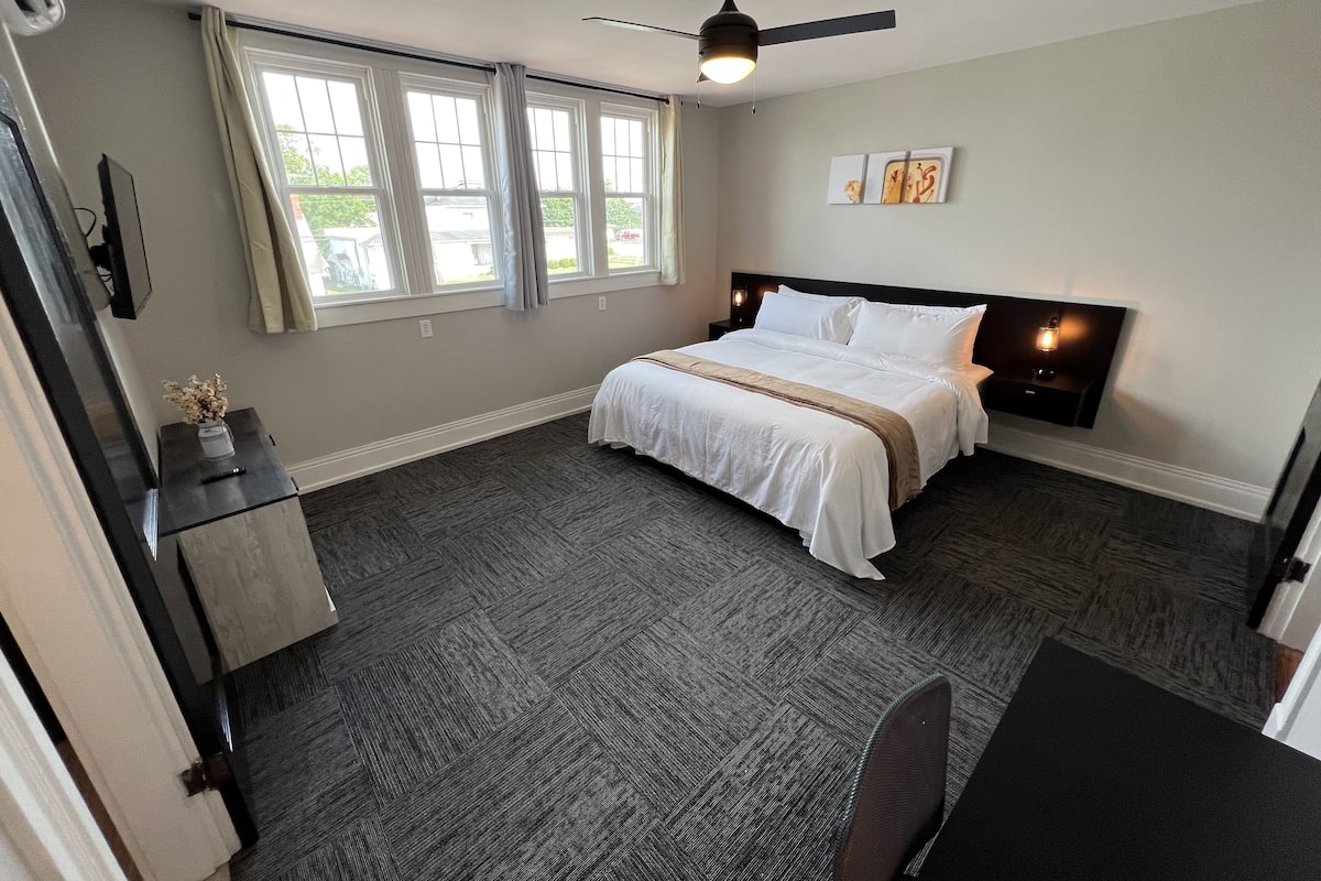Een slaapkamer met een bed, een bureau, een stoel, een plafondventilator en een raam met gordijnen.