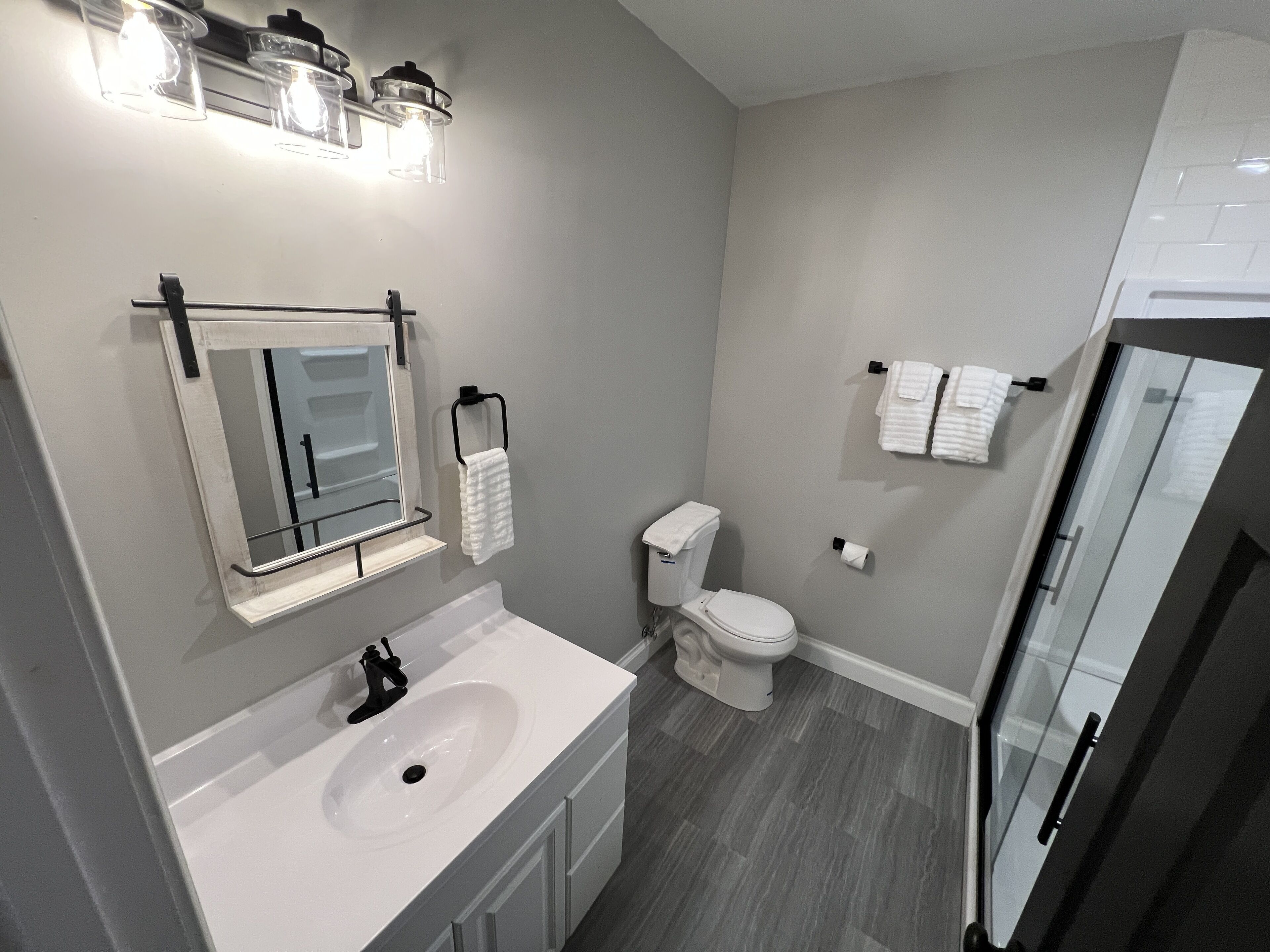 Un baño moderno con lavamanos, espejo y inodoro blancos. Hay varios toalleros y una puerta de cristal para la ducha.