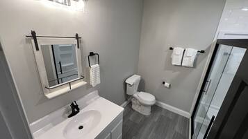 Un baño moderno con lavamanos, espejo y inodoro blancos. Hay varios toalleros y una puerta de cristal para la ducha.