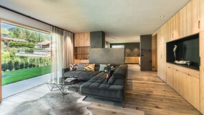 A modern living room with a sectional sofa, wooden flooring, and a large window offering a view of a green landscape. - 181 Amie Chalet (Santa Cristina Valgardena)