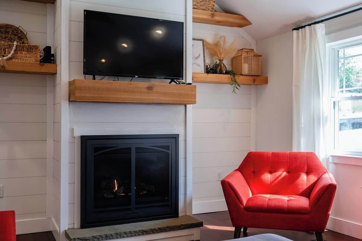 A living room with a flat-screen TV mounted above a fireplace, a red armchair, and wooden shelves with decorative items.