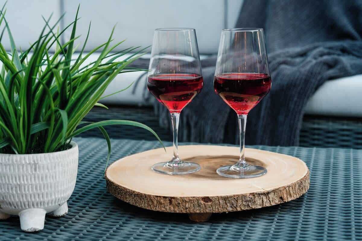 Deux verres de vin rouge posés sur un sous-verre en bois, une plante en pot et une surface texturée.