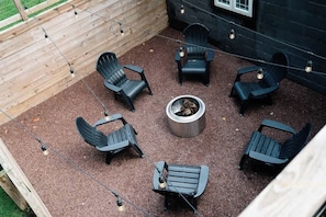 A patio with six Adirondack chairs arranged around a central fire pit, with string lights overhead.