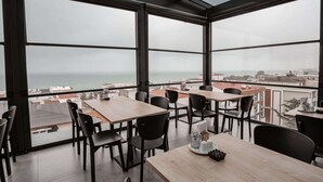 A modern restaurant with large windows offering a view of the sea and cityscape. Tables and chairs are arranged for dining. - Daric Hotels (Atakum)