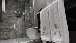 A bathroom with a marble wall, a glass-enclosed shower, and a white toilet. There are two towels hanging on the wall with the text "DARIC HOTELS". - Daric Hotels (Atakum)