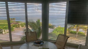 Una mesa redonda de vidrio con sillas, un tazón pequeño con frutas y un bloc de notas en un balcón con ventanas amplias y vistas a la vegetación y al agua.