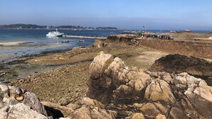 Un rivage rocheux avec un petit bateau blanc amarré près de l’eau, un ciel dégagé et une vue lointaine sur une ville.