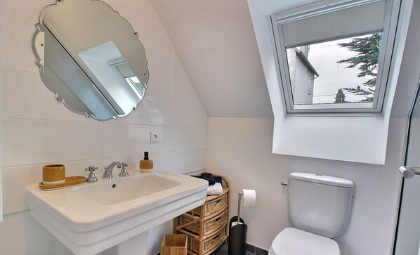 Une salle de bain moderne avec un lavabo blanc, un miroir et des toilettes. Il y a une fenêtre au-dessus des toilettes, un panier en osier et une petite poubelle.
