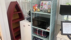 A compact room with a white refrigerator, a teal metal shelf unit with various items, a red storage unit, and a small desk with a computer monitor. - Great One Room with Full Bath Guest House! (Darien)