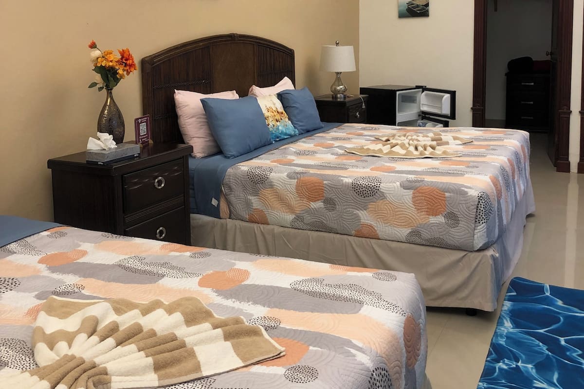 A hotel room with two beds, a nightstand with a lamp, a painting on the wall, and a door leading to another room.
