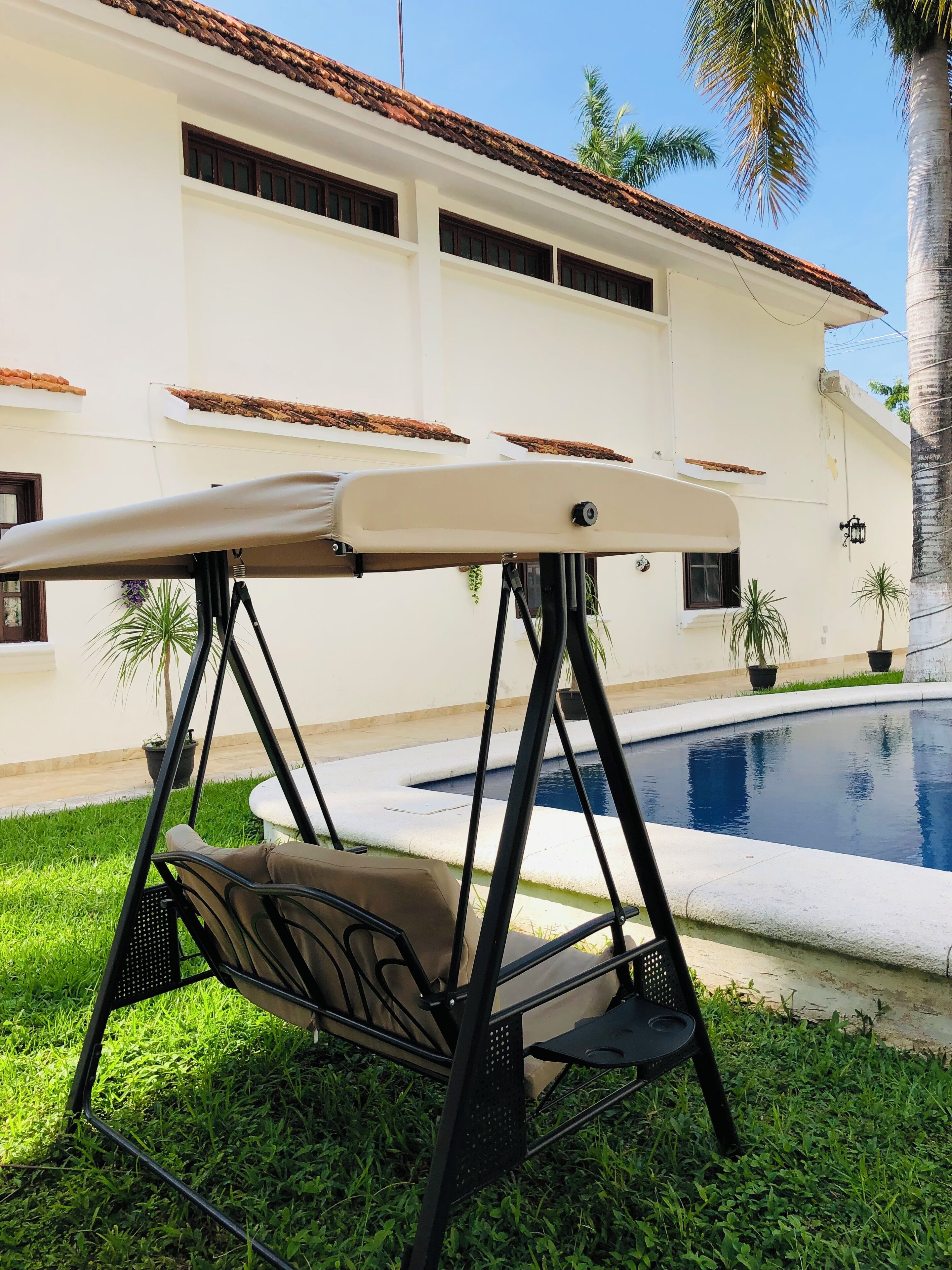A swing chair in a garden area with a pool and a white building in the background.