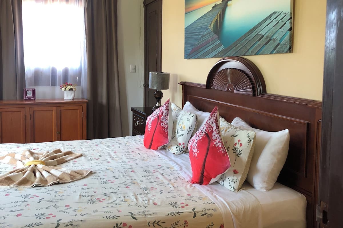 A hotel room with a large bed, a painting of a sunset over a pier, an air conditioning unit, and a wooden nightstand with a lamp.