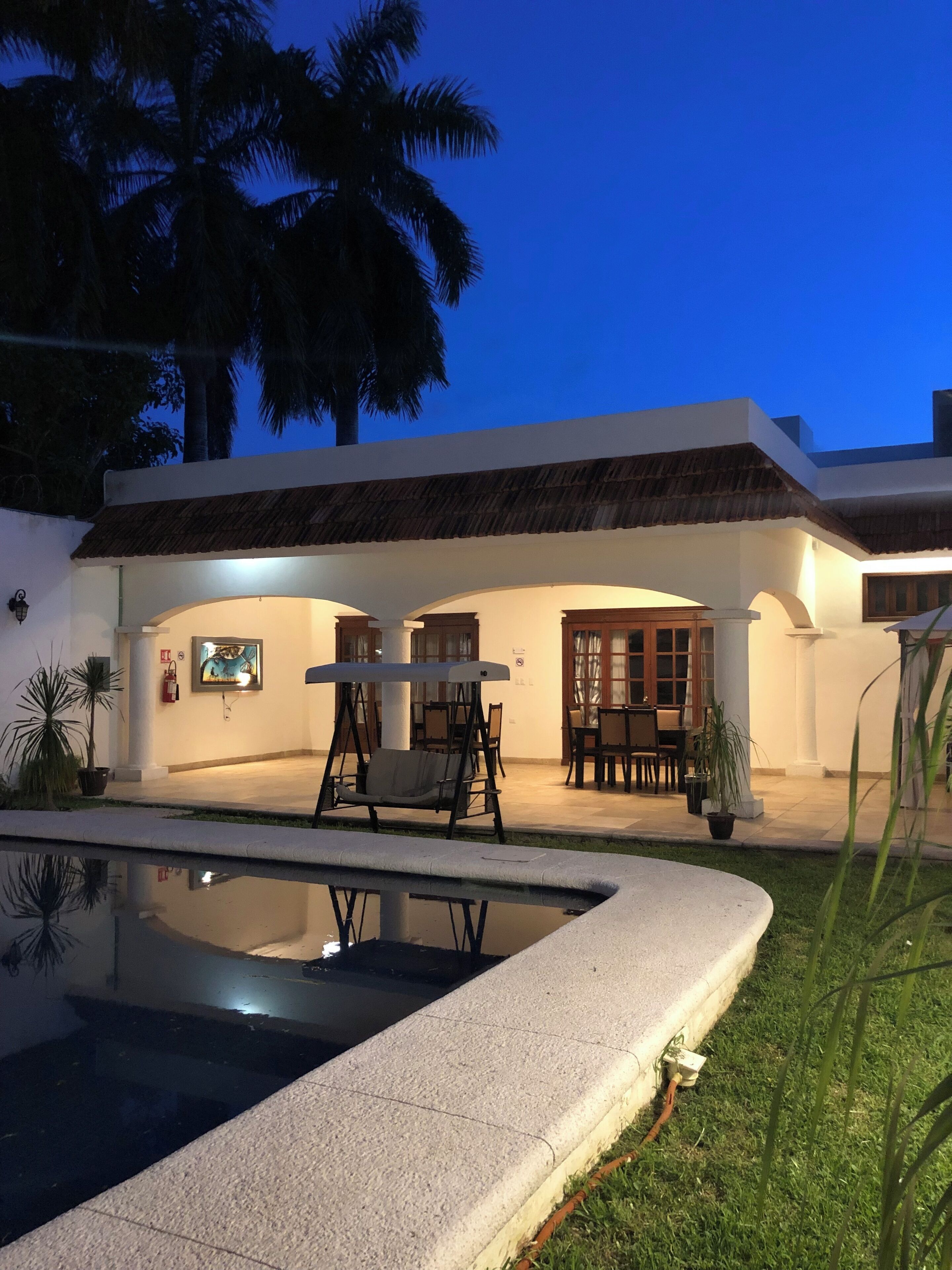 Una piscina con borde de piedra, mobiliario exterior y un edificio con techo de paja y paredes blancas.