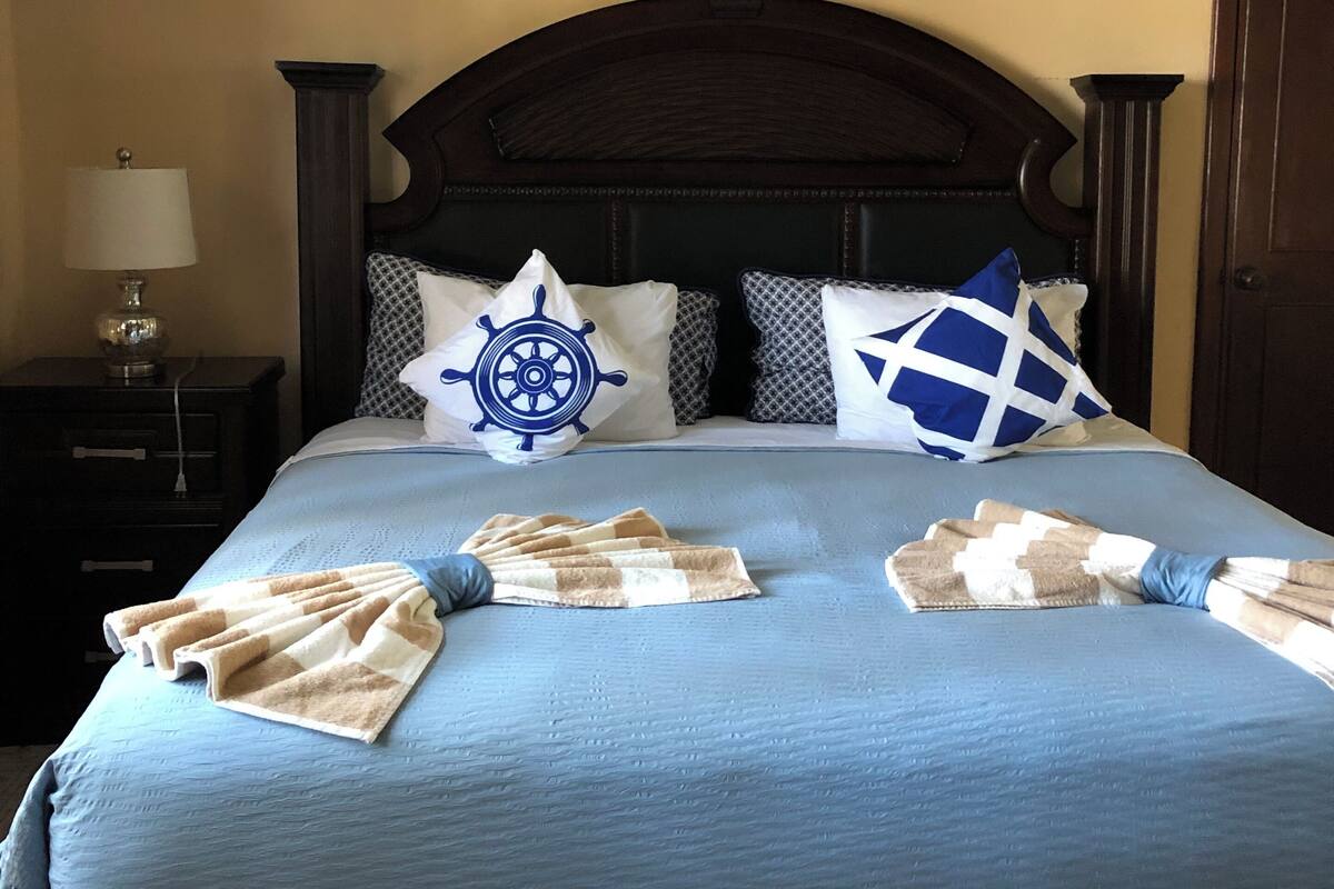 A neatly made bed with a blue bedspread, a wooden headboard, and decorative pillows including a ship's wheel and a blue and white checkered pillow.