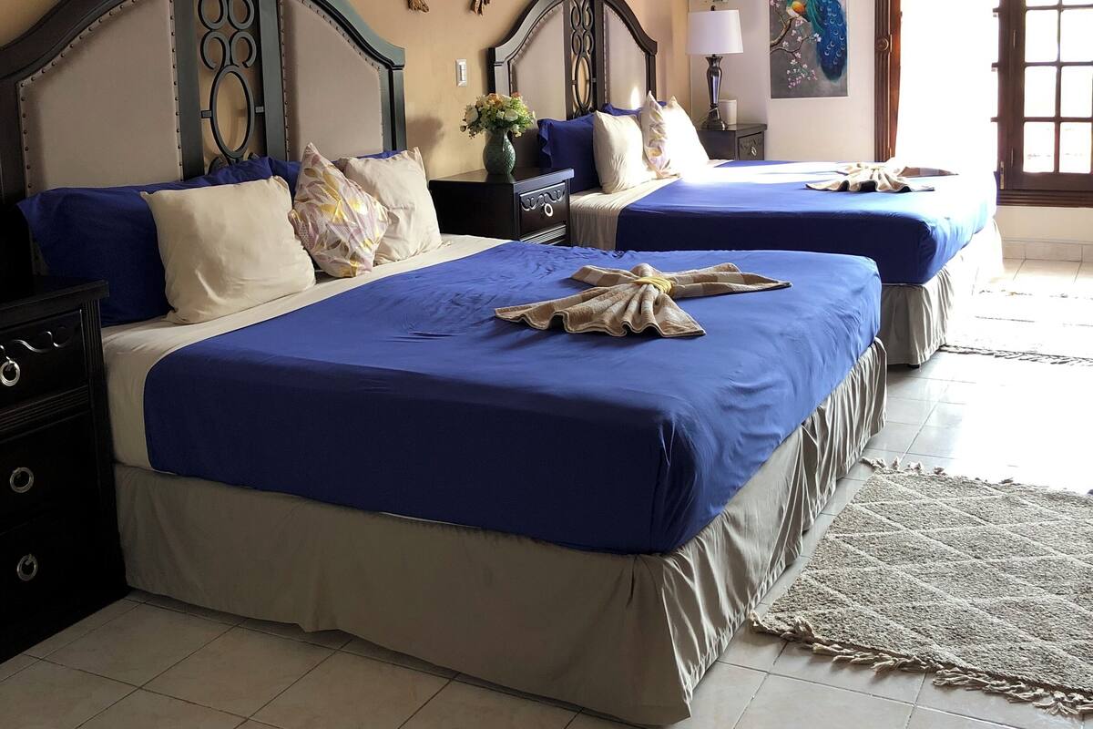 A hotel room with two beds, a ceiling fan, a decorative wall piece, and a window with curtains.