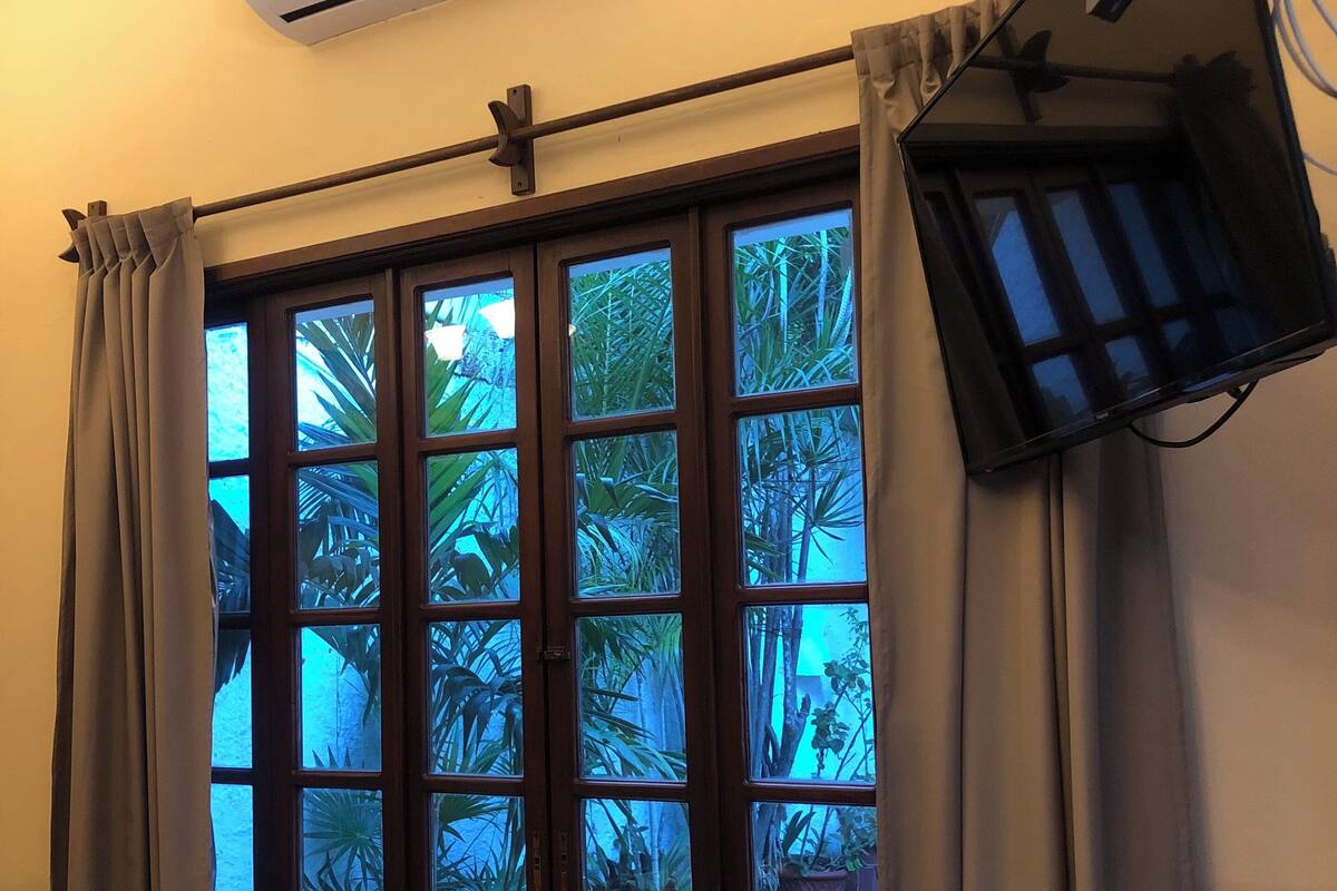 A room with a window, curtains, an air conditioner, a flat-screen TV mounted on the wall, and a small table with a decorative item and a sign.