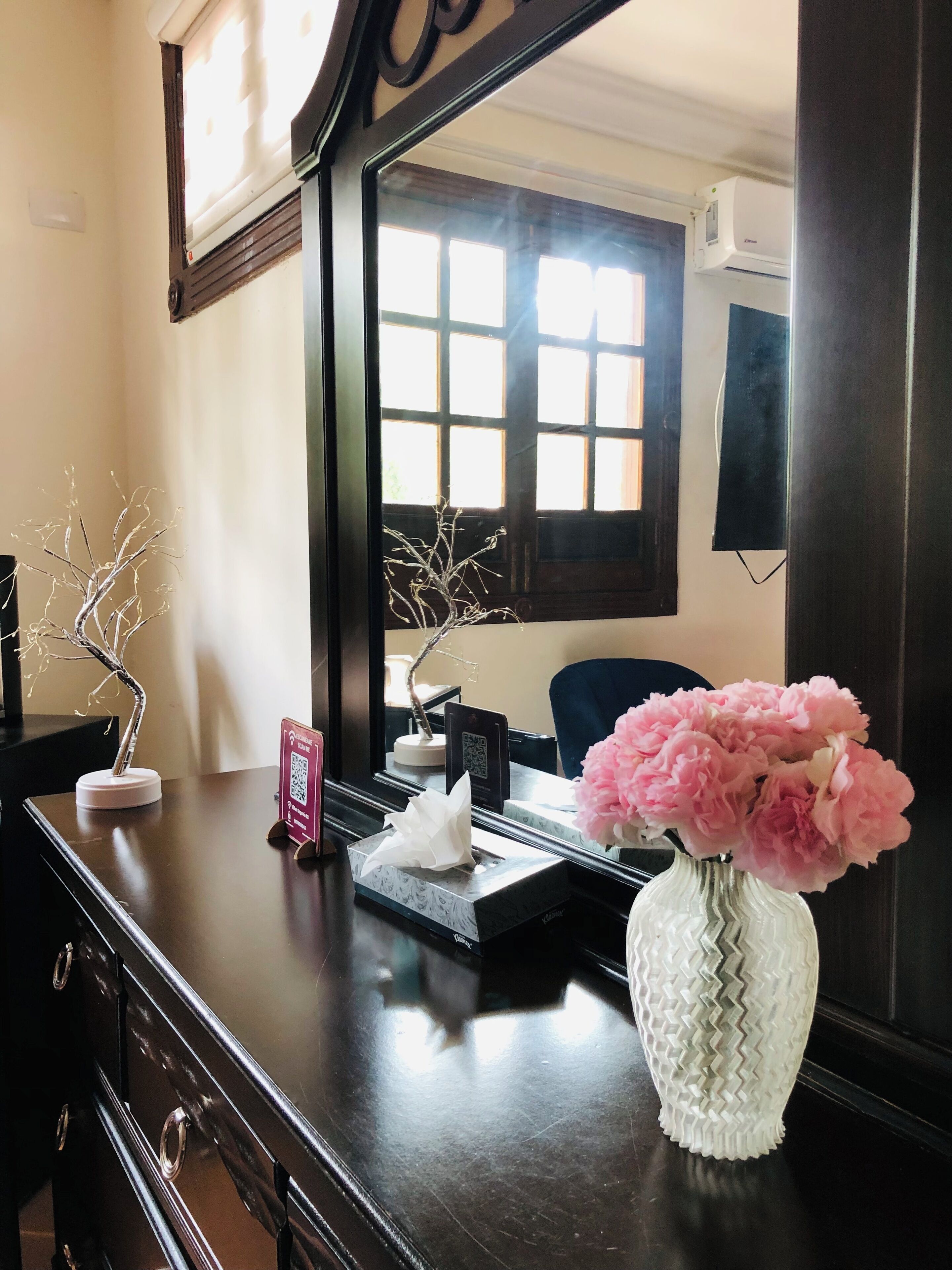 A hotel reception desk with a mirror, a vase of pink flowers, a tissue box, a QR code sign, and a decorative tree.