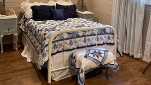 Un dormitorio con techo de madera, una cama con colcha floral, dos mesitas de noche con lámparas, una ventana con diseño de vitral y una pared con cuadros enmarcados.