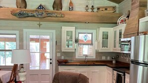 Un salón espacioso con un sillón marrón, una mesa redonda y un cojín decorativo. Se ve una cocina en el fondo con armarios blancos y electrodomésticos de acero inoxidable.