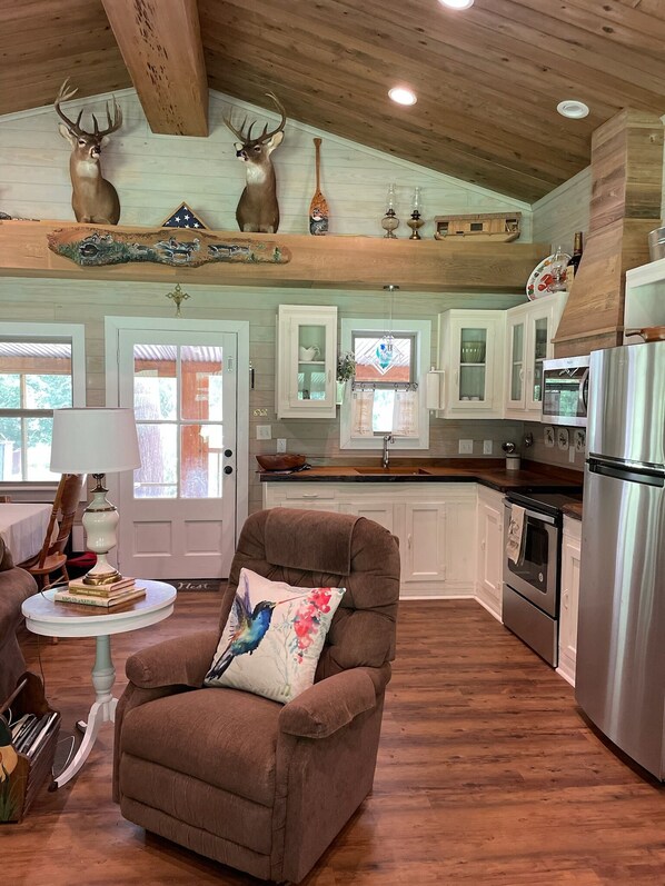A spacious living room with a brown armchair, a round table, and a decorative pillow. A kitchen with white cabinets and stainless steel appliances is visible in the background.