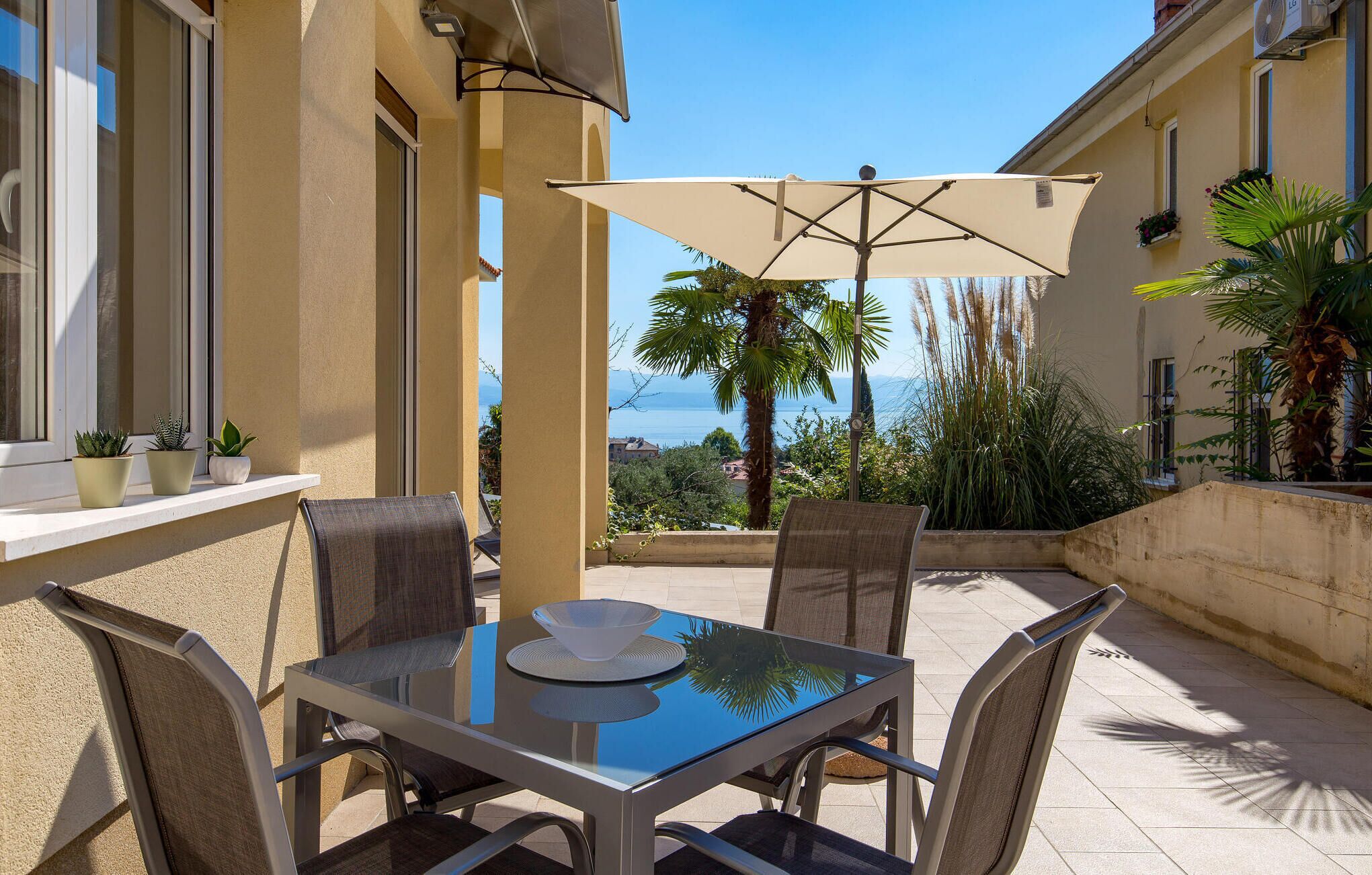 A glass-top table with four chairs and a beige umbrella in a sunlit outdoor area with a view of the sea.