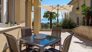 A glass-top table with four chairs and a beige umbrella in a sunlit outdoor area with a view of the sea. - 1 bedroom awesome apartment in Lovran (Lovran)