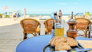 Ein Tisch mit einer Flasche Blanquette de Naumer, einem Glas Saft und Brotscheiben in einem sonnigen Café direkt am Strand.