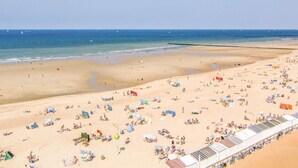 Ein Strand mit vielen Liegestühlen und Sonnenschirmen, ein sandiger Uferstreifen und ein ruhiges Meer mit Booten in der Ferne.