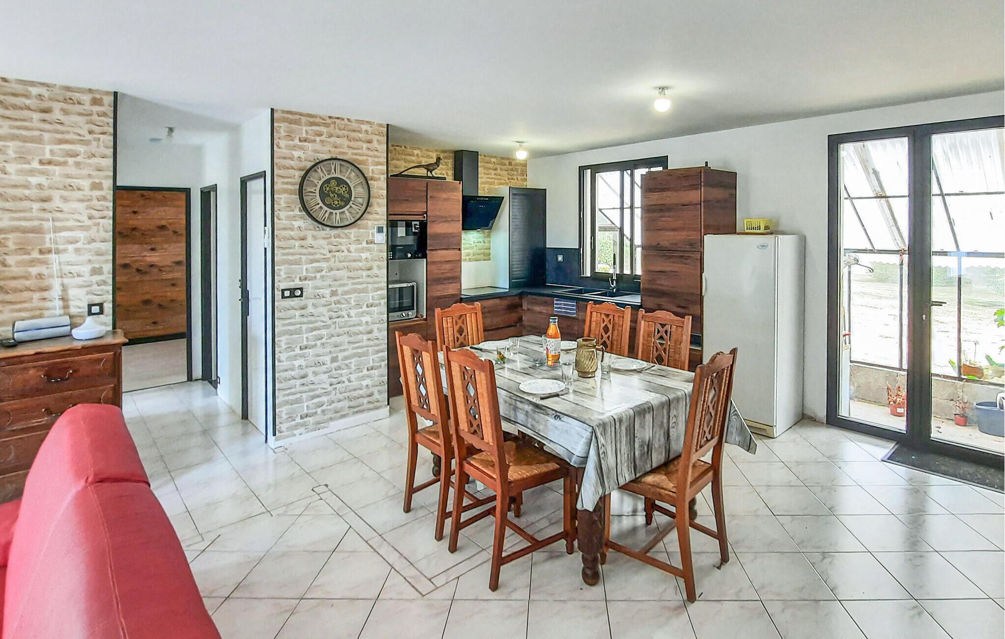 A dining area with a table set for four, surrounded by wooden chairs. A red sofa is visible in the foreground. The room features a brick wall with a decorative clock, a wooden cabinet, and a large window with a view of the outdoors.