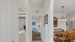 Corredor que dá para um quarto com cama, um banheiro com chuveiro e uma área de jantar com mesa e cadeiras.