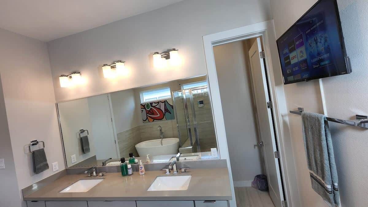 Een moderne badkamer met een grote spiegel, twee wastafels, een douche en een aan de muur bevestigde televisie.
