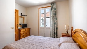 Une chambre à coucher comprenant un lit, une commode, un téléviseur, une fenêtre avec des rideaux et une table de chevet avec une lampe.