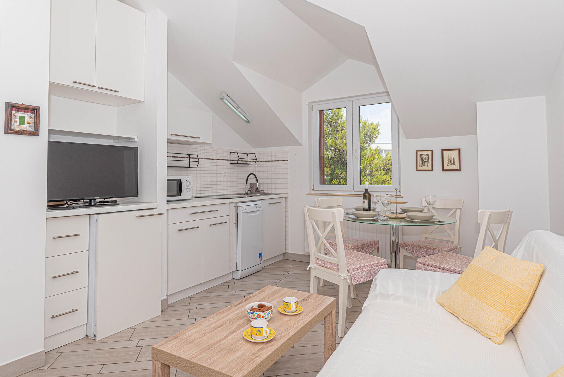 A compact kitchen with white cabinets, a microwave, and a stove. There is a dining area with a glass table, white chairs, and a sofa with a yellow pillow.