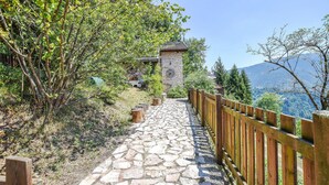 Ein steinbelagter Weg führt zu einem Steinhaus mit einer Uhr, umgeben von Grünpflanzen und einem Holzzaun.