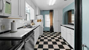 Una cucina con mobili bianchi, pavimento a scacchi bianconero e una finestra con tende.