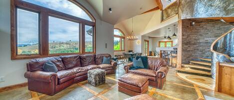 A spacious living room with leather sofas, a stone fireplace, and large windows offering a view of the landscape.