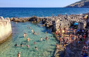 Una playa rocosa con aguas cristalinas y gente nadando y tomando el sol.